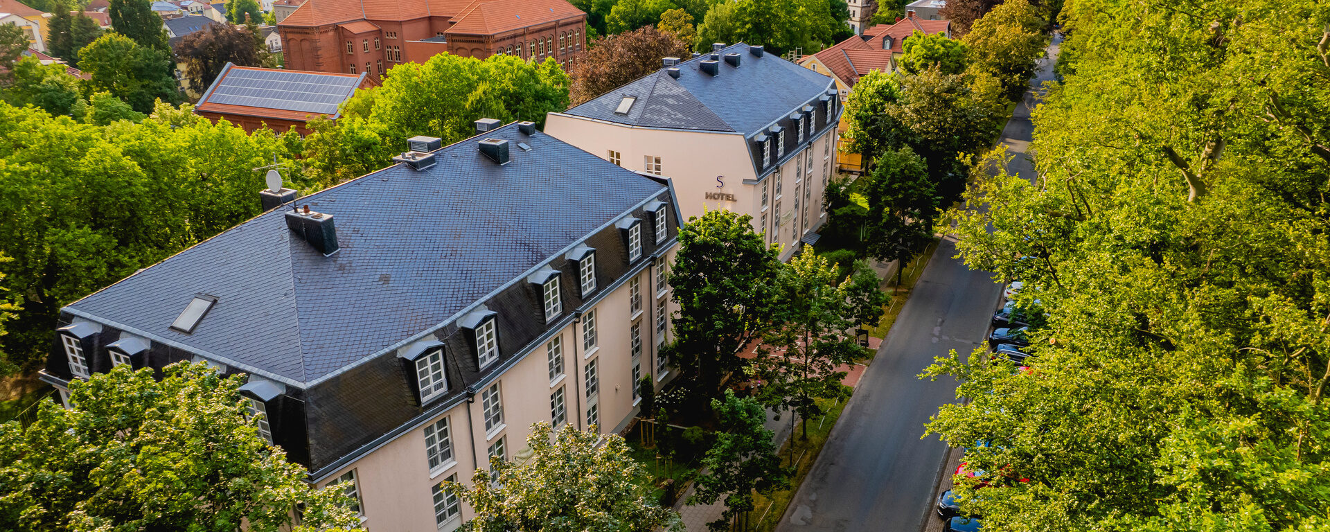Das Bild zeigt das Hotel am Schosspark von oben umgeben von gr&uuml;nen B&auml;umen ´